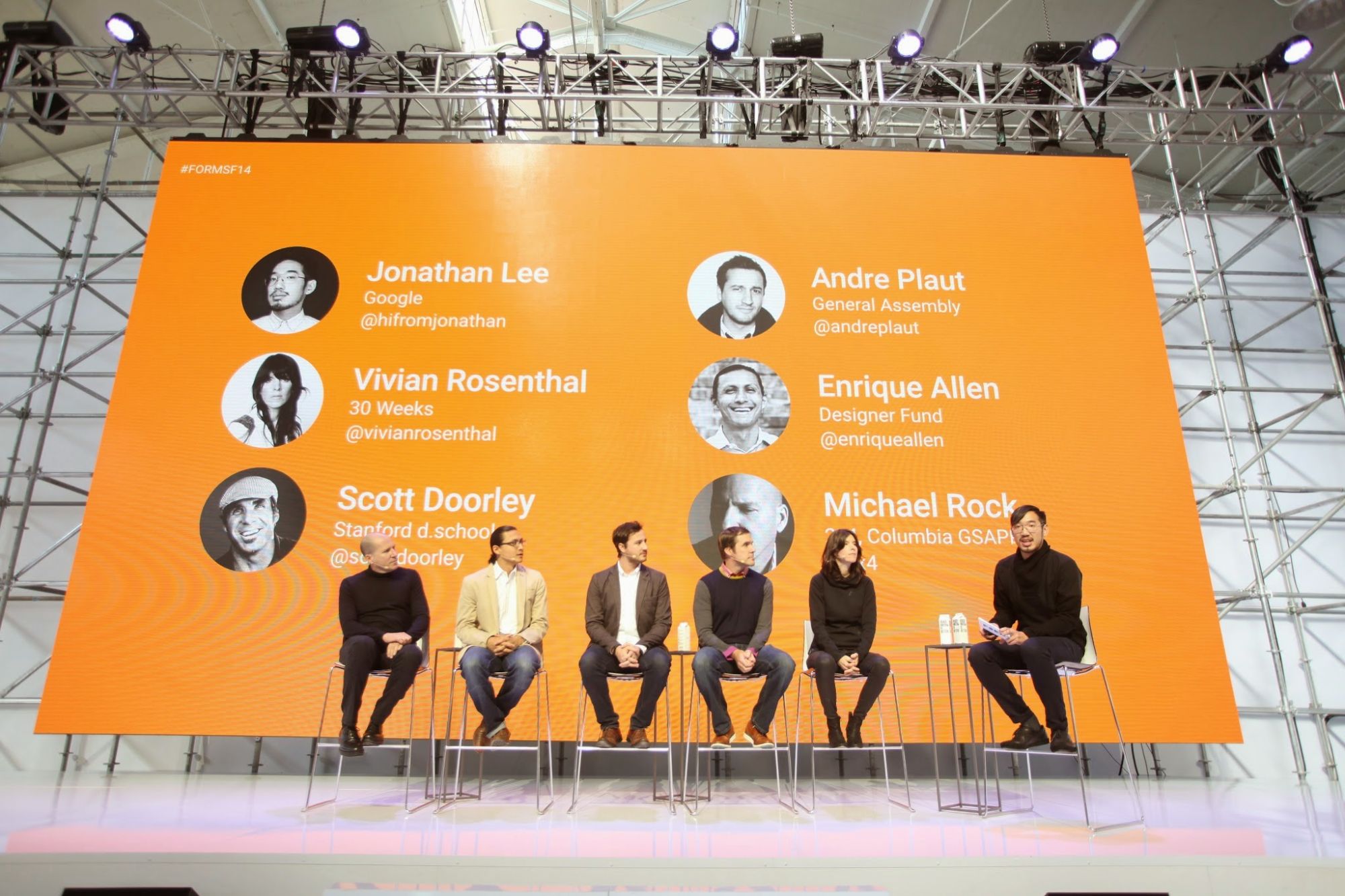 Six panelists seated on stools on stage in front of an orange screen listing their names