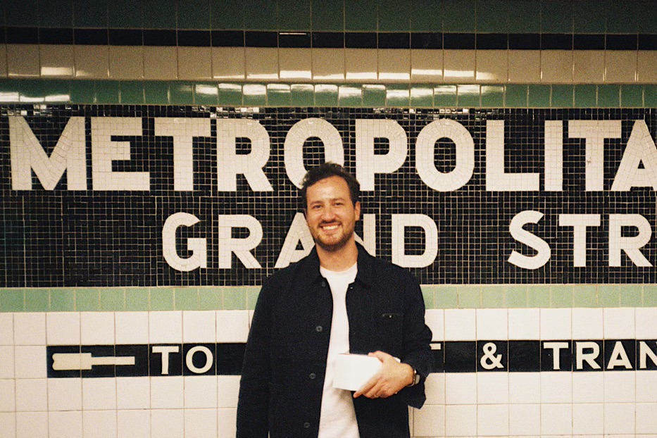 Andre smiling in front of a tiled subway sign reading Metropolitan and Grand Street